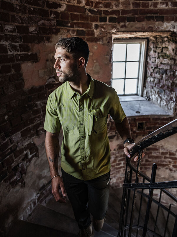 A man in a green Halti Samoy Trekking Shirt stands on a spiral staircase by a window in a brick building, looking left.