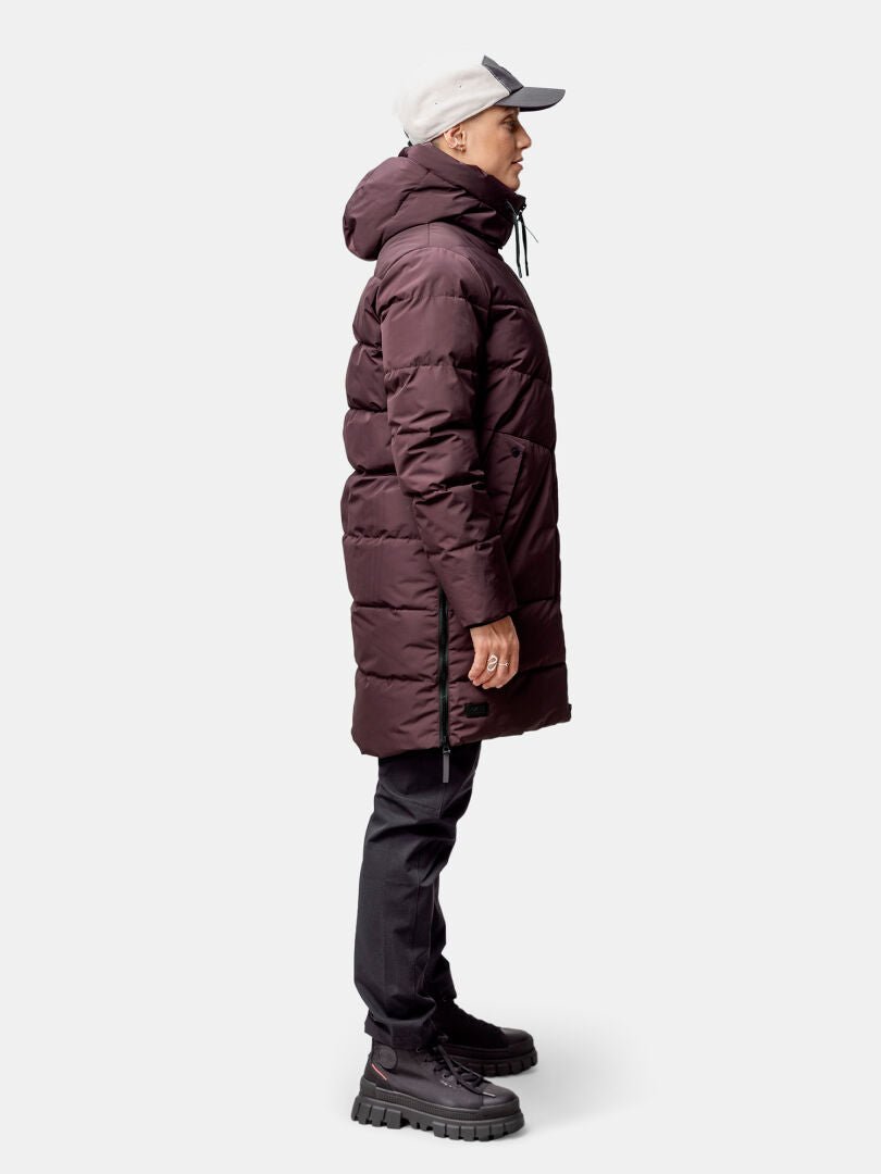 A woman in a Halti Linjat Parka Jacket, black pants, boots, and a light cap stands in profile against a plain background.