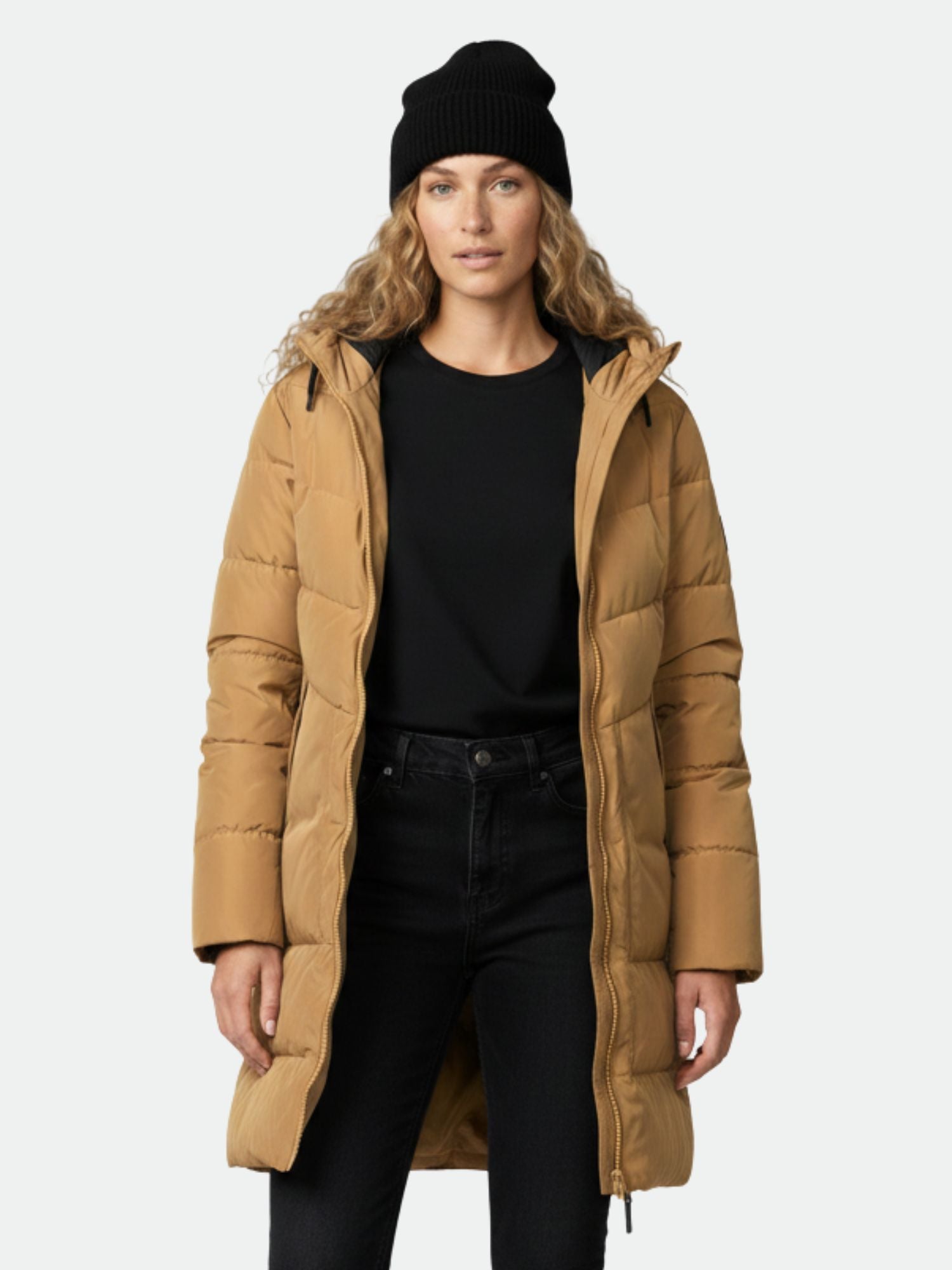 A woman in a tan Halti Linjat Parka Jacket, black beanie, shirt, and pants stands facing forward against a plain background.