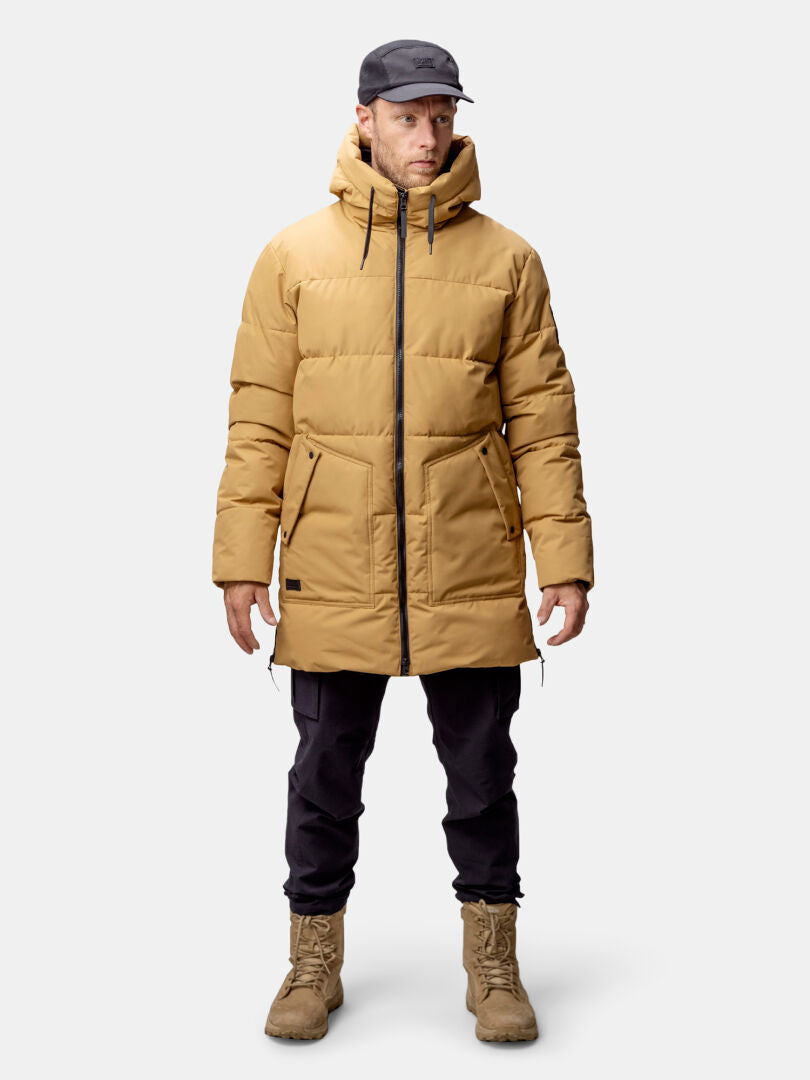 A man in a tan Halti Linjat Parka Jacket, black pants, tan boots, and a black cap stands against a plain background.
