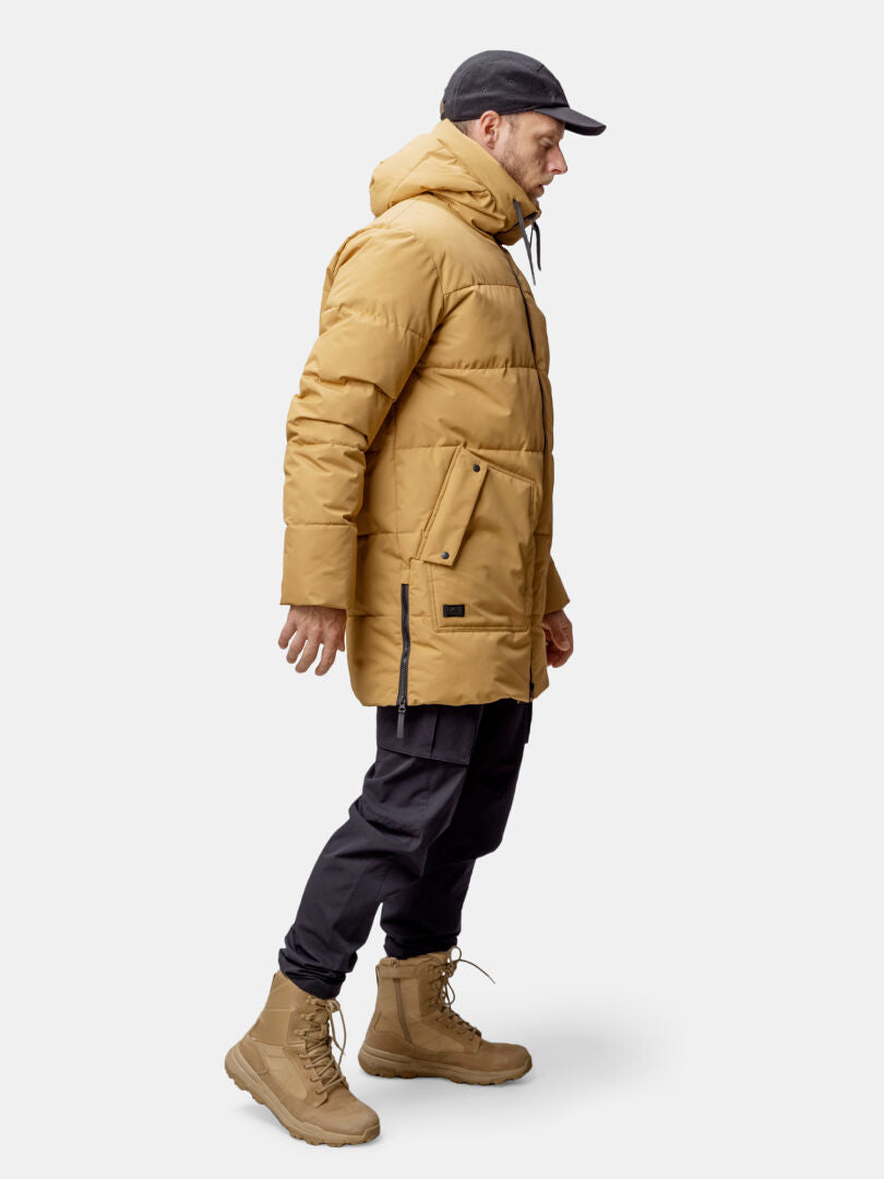 A man wears the Halti Linjat Parka Jacket, black pants, tan boots, and a black cap against a plain light background.