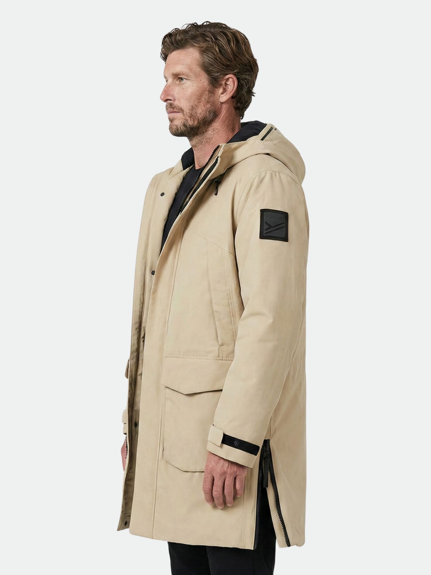 A man in a beige Halti Bergga DX Winter Parka with front pockets stands facing left against a plain background.