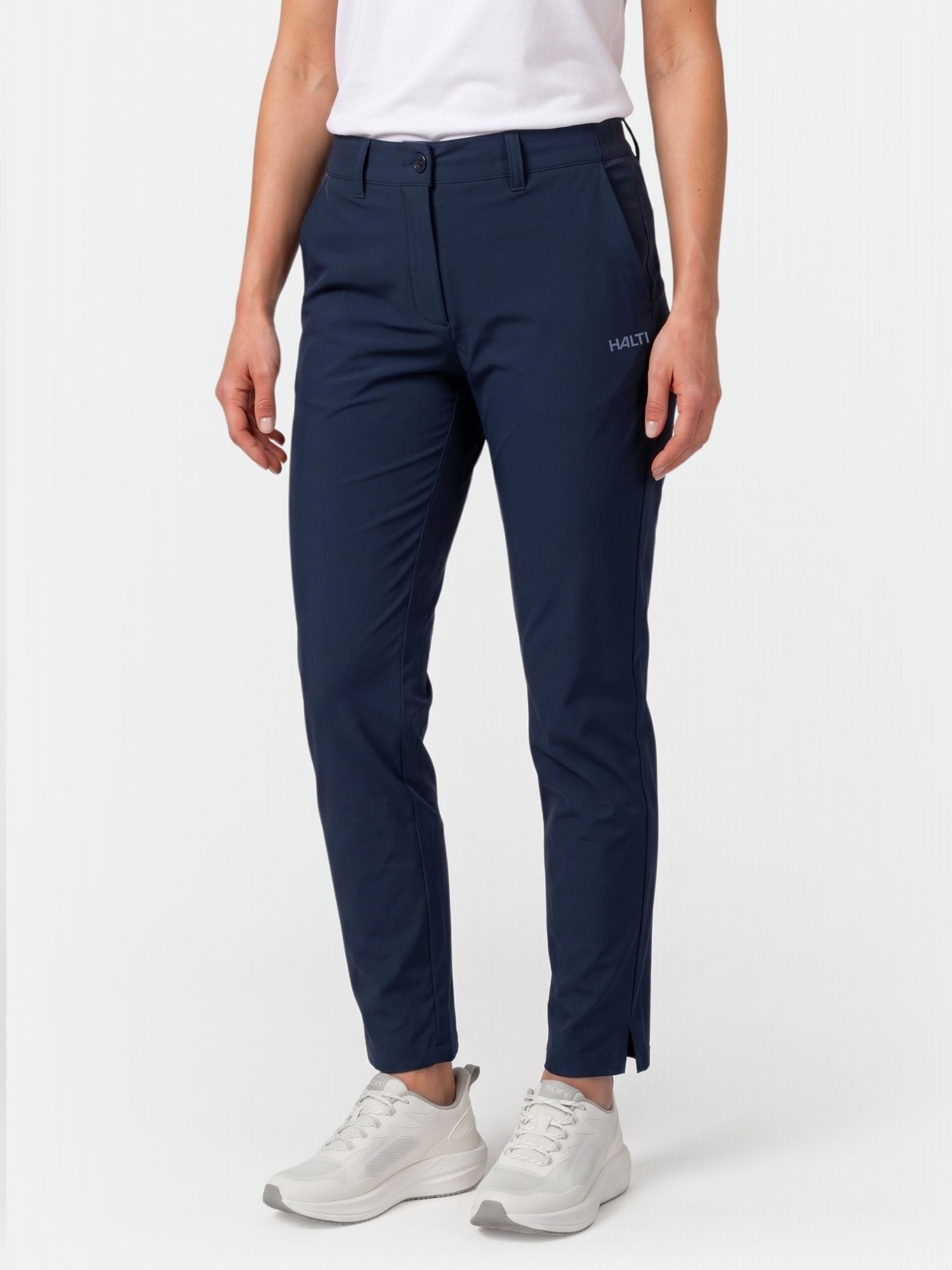 Woman in Halti Drive X-stretch Chinos, white tee, and sneakers stands against a plain background.