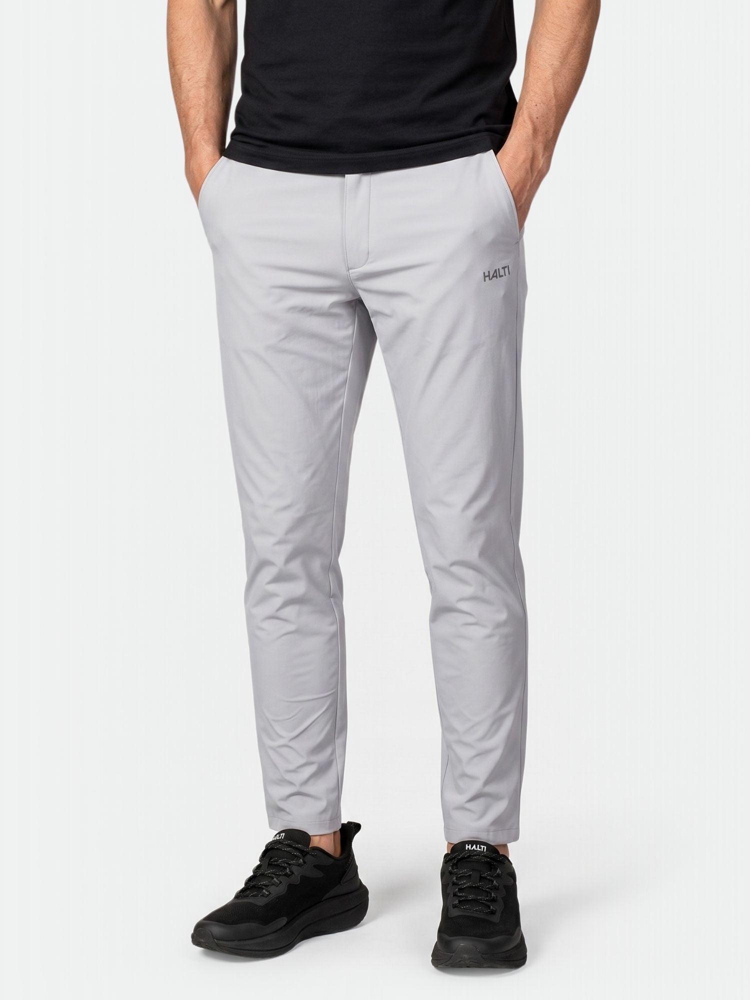 A person in Halti Drive X-stretch Chinos, a black shirt, and black sneakers stands hands-in-pockets against a plain background.