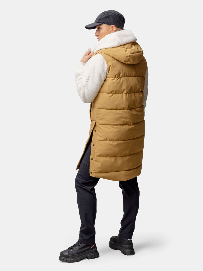 A woman faces away in a tan Halti Penger Puffer Vest with hood, white fleece sleeves, black pants, and boots.