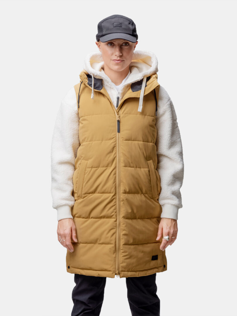 Woman in Halti Penger Puffer Vest, tan with hood, over white hoodie, black pants, and black cap.
