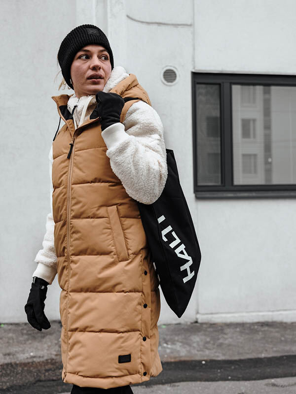 Woman in a tan Halti Penger Puffer Vest and beanie carries a tote, walking past a white building.