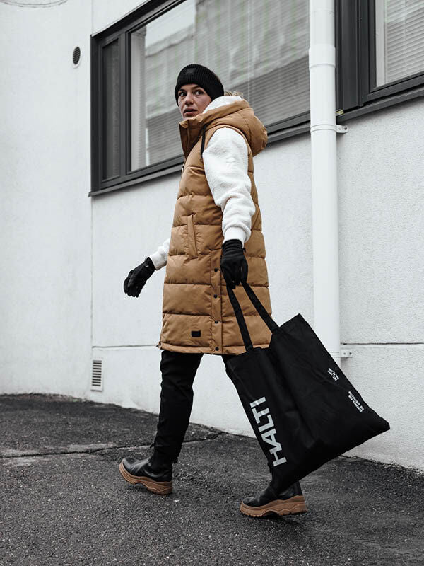 A person in a tan Halti Penger Puffer Vest and black beanie walks, carrying a large black Halti tote bag.