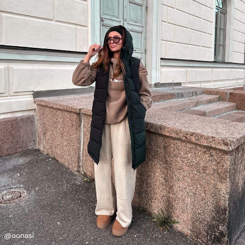 A woman in Halti's Penger Puffer Vest and sunglasses stands on a sidewalk by a stone staircase and building wall.