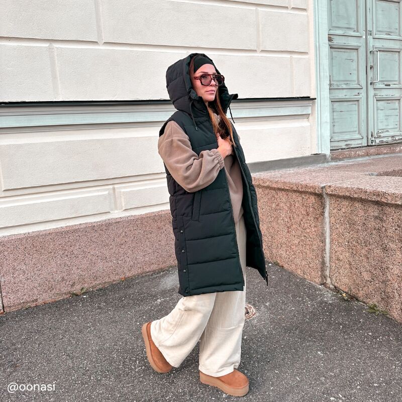 Woman wears Halti Penger Puffer Vest, beige sweater, wide pants, and boots while walking by a stone wall in the city.