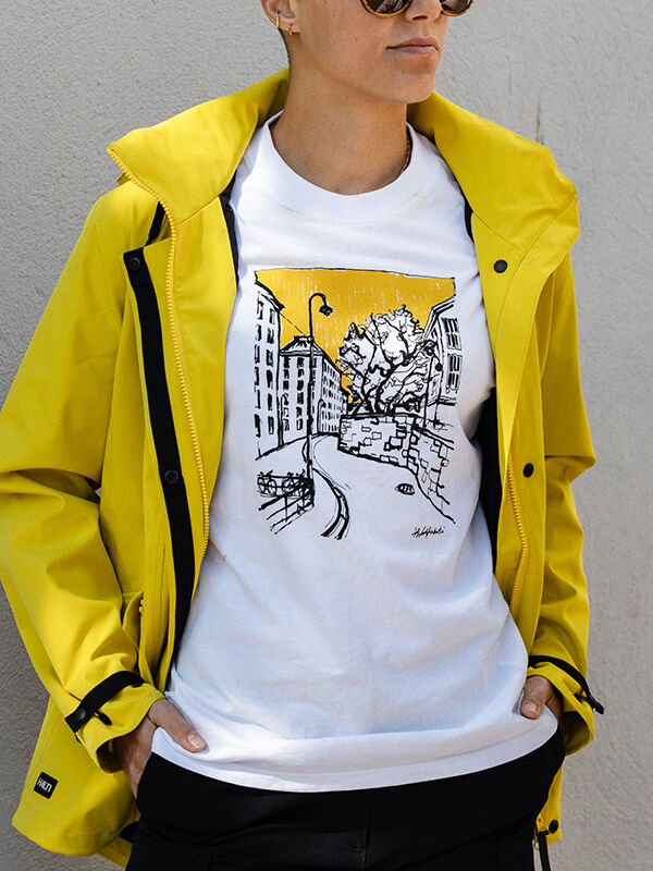 Person in sunglasses, yellow jacket, and Halti Skaltsi Unisex T-shirt stands against a gray wall.