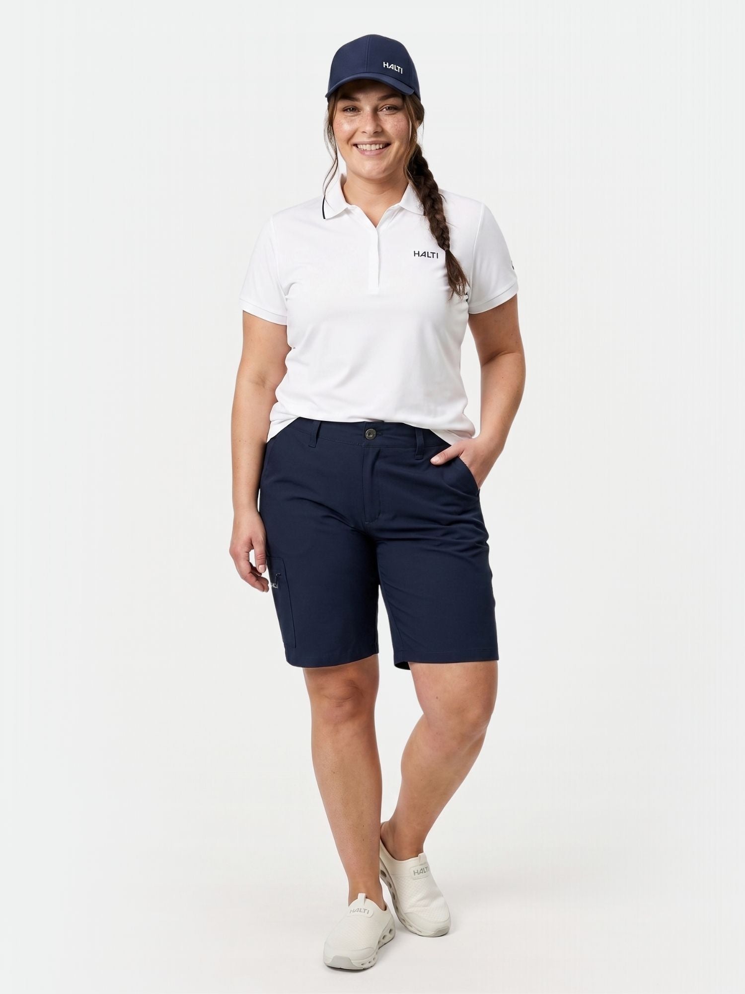 Woman wearing Halti Drive X-stretch Shorts Women's Plus, navy cap, and white polo stands against a white background.