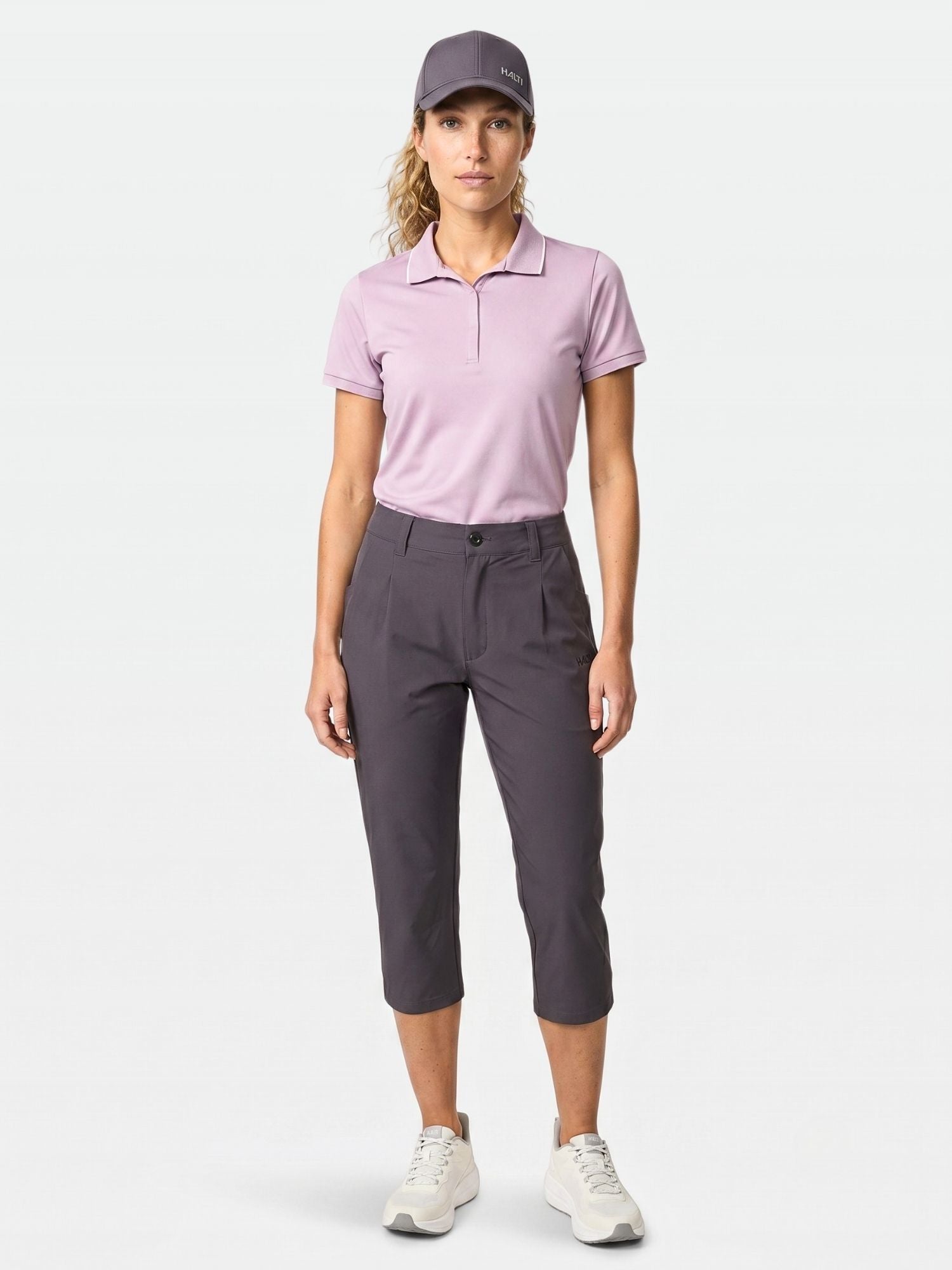 A person stands facing forward in a UPF 50+ lavender polo, Halti Drive X-stretch capri pants, gray cap, and white sneakers.