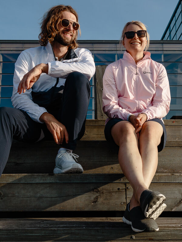 A man and woman in sunglasses smile outdoors, both wearing Halti Saime Windbreaker Jackets on wooden steps.