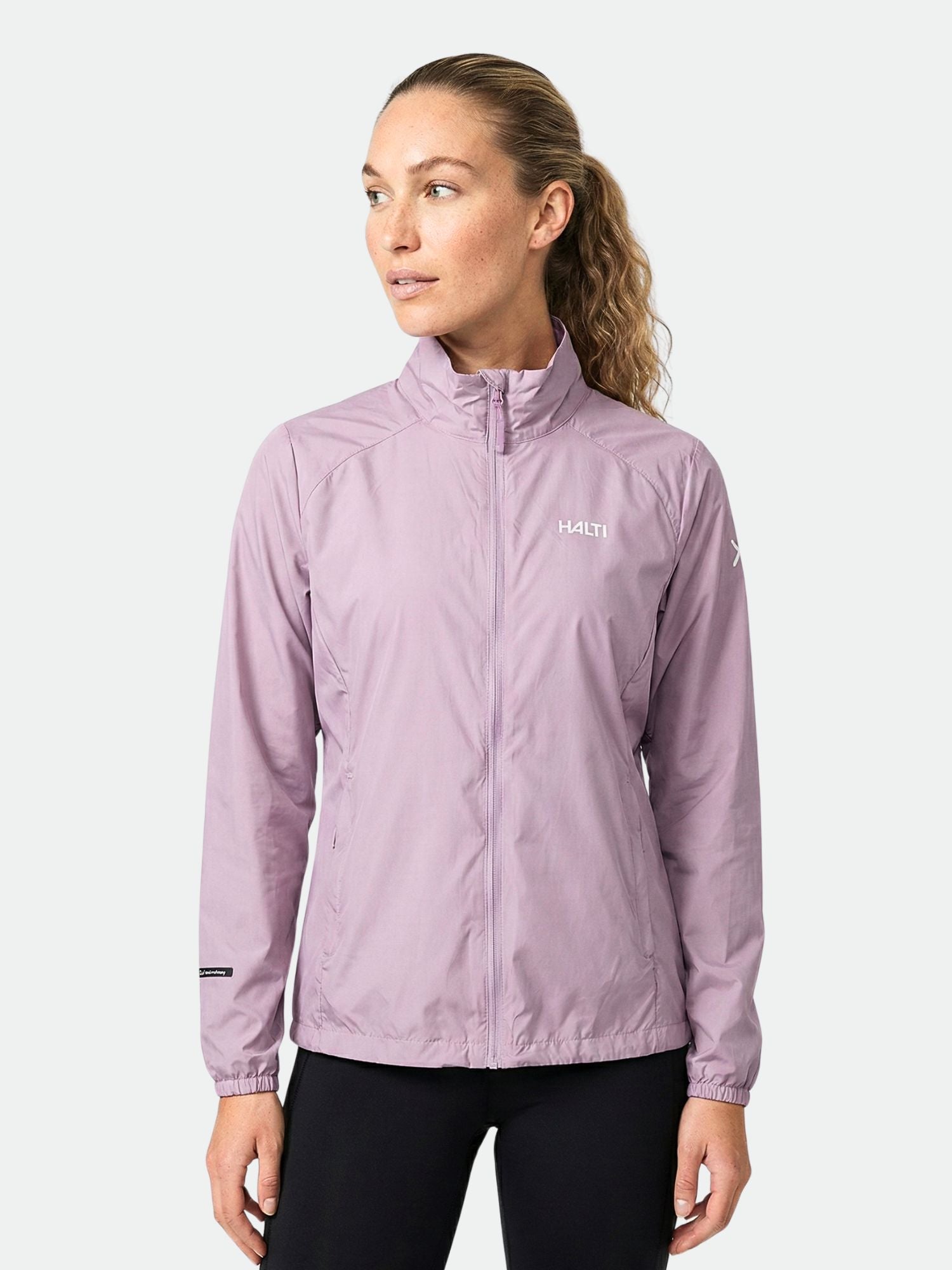 A woman wears the Halti Saime Windbreaker Jacket in light purple with black pants, standing against a plain background.