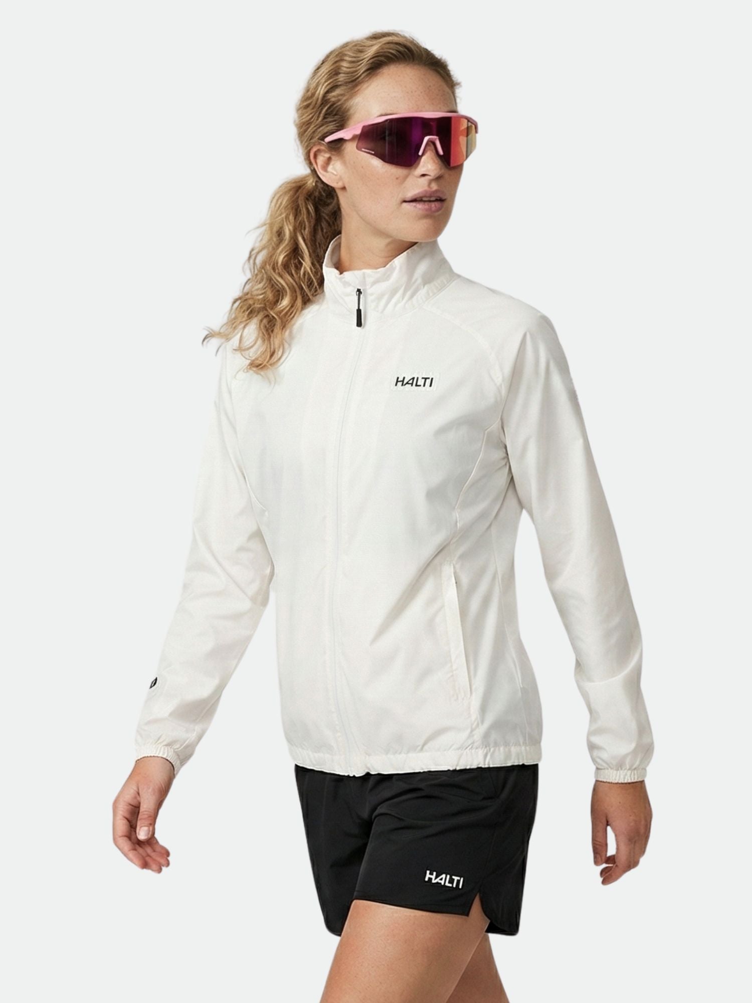 A woman in the Halti Saime Windbreaker Jacket poses confidently in black shorts and red sunglasses.