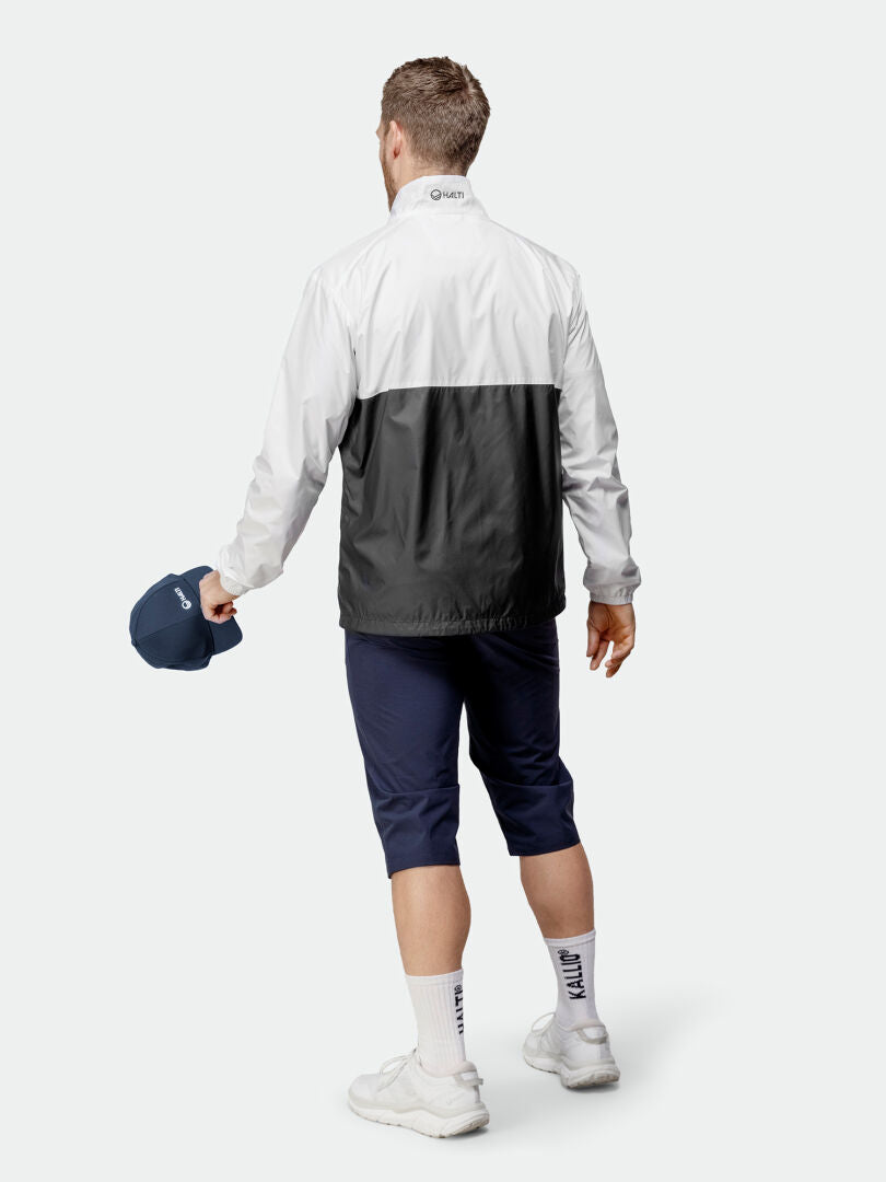 A man in the Halti Saime Windbreaker Jacket, navy pants, white socks and shoes, holds a navy cap.