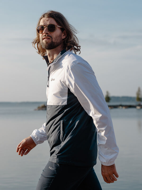 A man in sunglasses wears the Halti Saime Windbreaker Jacket Men's near calm water under a clear sky.