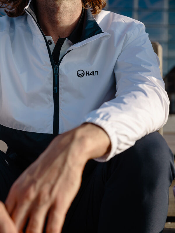 Man wearing a Halti Saime Windbreaker Jacket (white/black, water-repellent) sits outdoors with arm on knee.