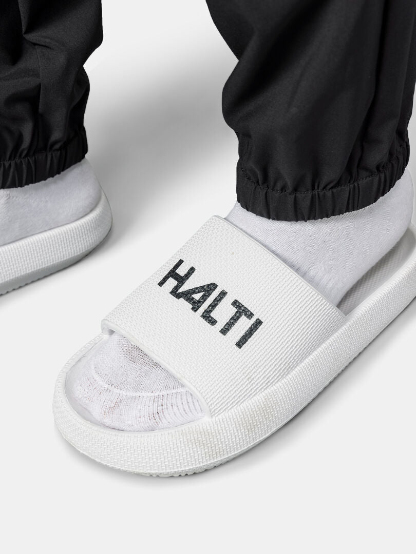 A person in Halti Grets Jogger Pants, white socks, and HALTI slides stands on a light surface.