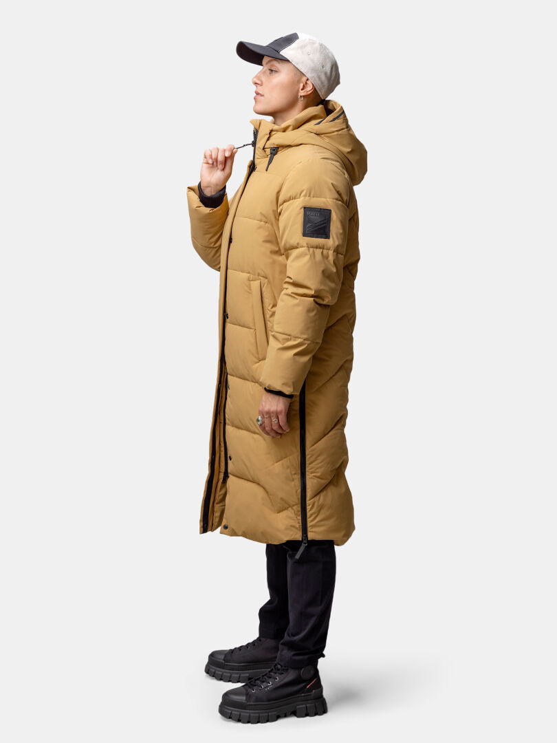 A woman in a Halti Brahe Puffer Long Winter Jacket, black pants, and a two-tone cap stands sideways.