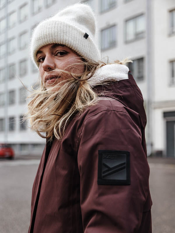 A woman in a maroon Halti Tokoi II DX Parka Jacket and white knit hat stands on a city street, facing the camera.