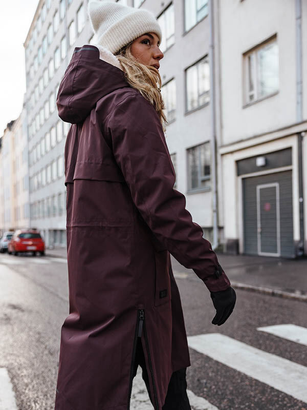 A woman in a maroon Halti Tokoi II DX Parka Jacket and white hat walks on a city street, glancing over her shoulder.