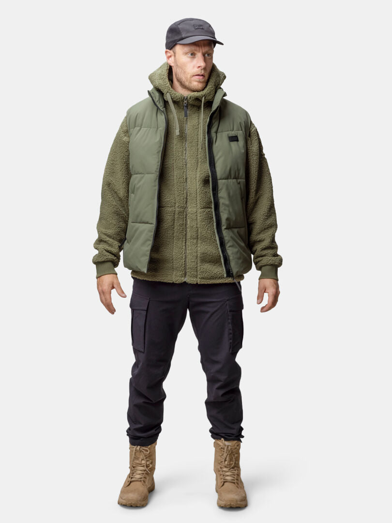 A man in a Halti Penger Classic Puffer Vest, fleece jacket, black pants, tan boots, and a black cap stands against a plain background.