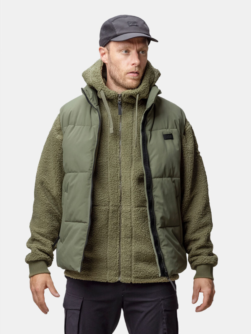 A man wears the Halti Penger Classic Puffer Vest, a green fleece hoodie, black cap, and cargo pants facing forward.