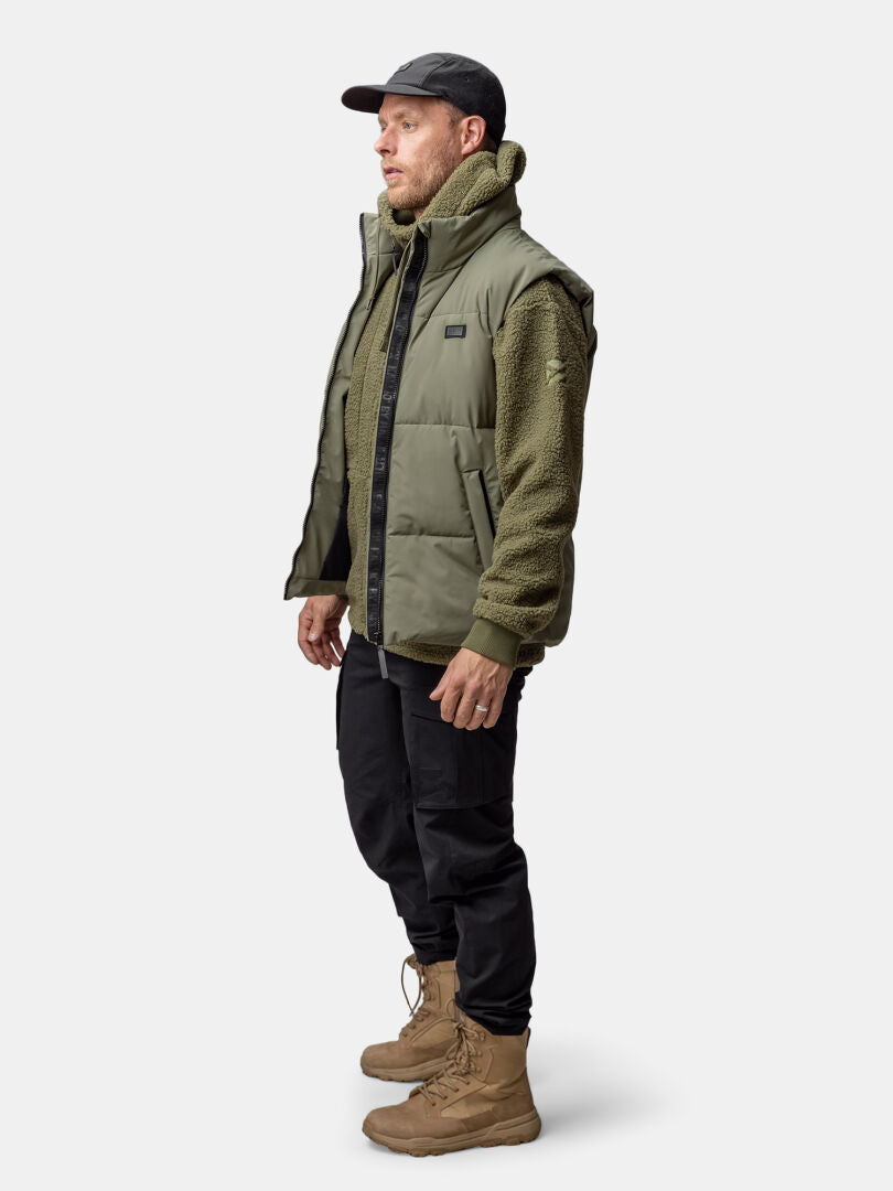 Man in Halti Penger Classic Puffer Vest, green fleece, black cap and pants, tan boots, standing sideways.