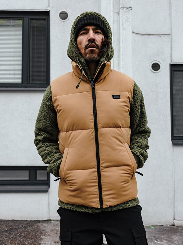 Man wears Halti Penger Classic Puffer Vest in tan over a green hooded jacket, standing outside by a white building.