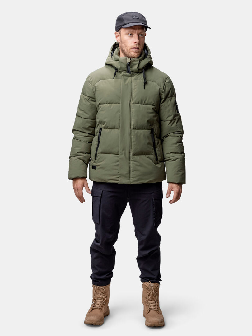 A man in a Halti Brahe Puffer Winter Jacket, black cargo pants, tan boots, and a black cap stands against a plain background.