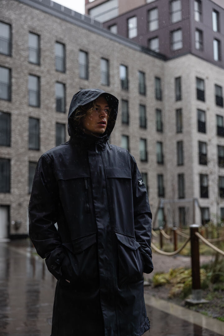Man wears Halti Tokoi II DX Parka Jacket, hands in pockets, outside a modern apartment on a rainy day.