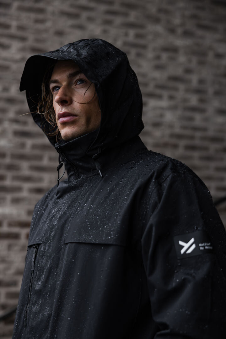 A man wears the Halti Tokoi II DX Parka Jacket, black and waterproof, standing outside by a brick wall in the rain.