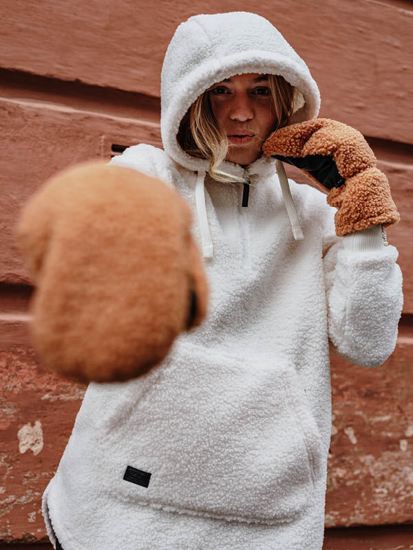 Person in a Halti Lykky Fleece Pullover poses with fists raised, wearing brown mittens by a reddish-brown wall.