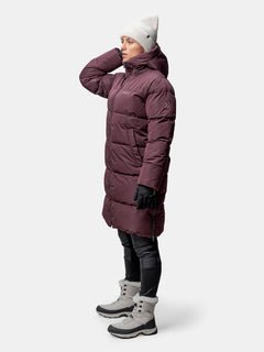 Wearing a Halti Sammal Insulated Parka Jacket, black gloves, winter boots, and a white beanie, she faces left.