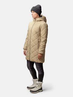 A woman stands sideways wearing the Halti Haapa Quilted Coat Jacket, black beanie, black pants, and beige winter boots.