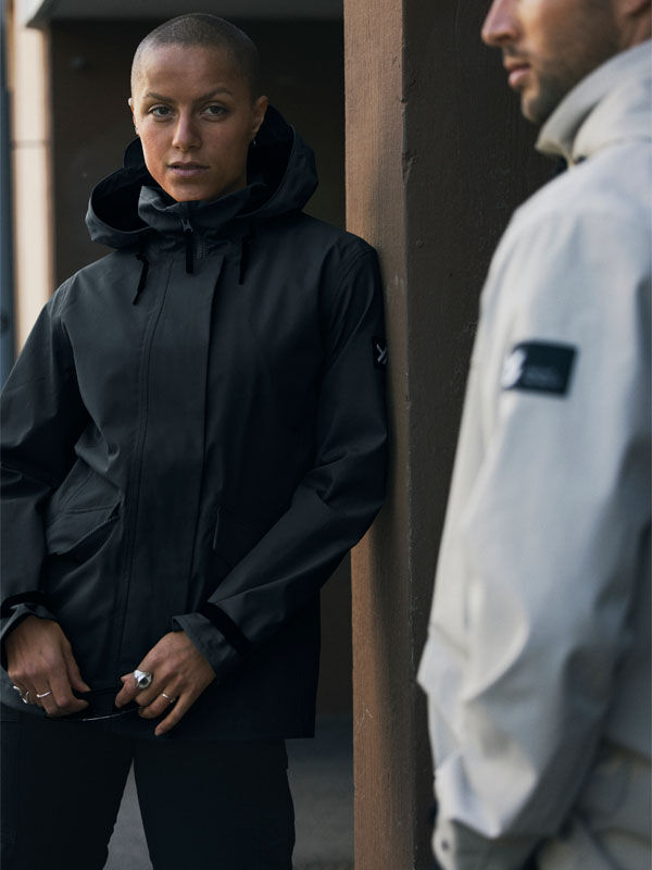A woman in a Halti Tokoi DX Jacket stands against a wall while another woman in a light jacket is in the foreground.