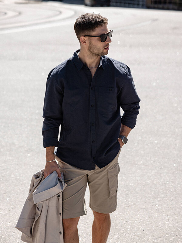 A man in a Halti Notsgi Longsleeve Shirt, khaki shorts, and sunglasses holds a light jacket on a sunny street.