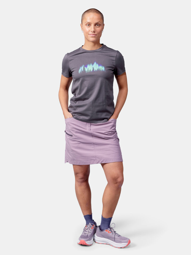 Person standing with hands in pockets, wearing Halti Matka II T-shirt Women's, mauve skirt, blue socks, gray sneakers.