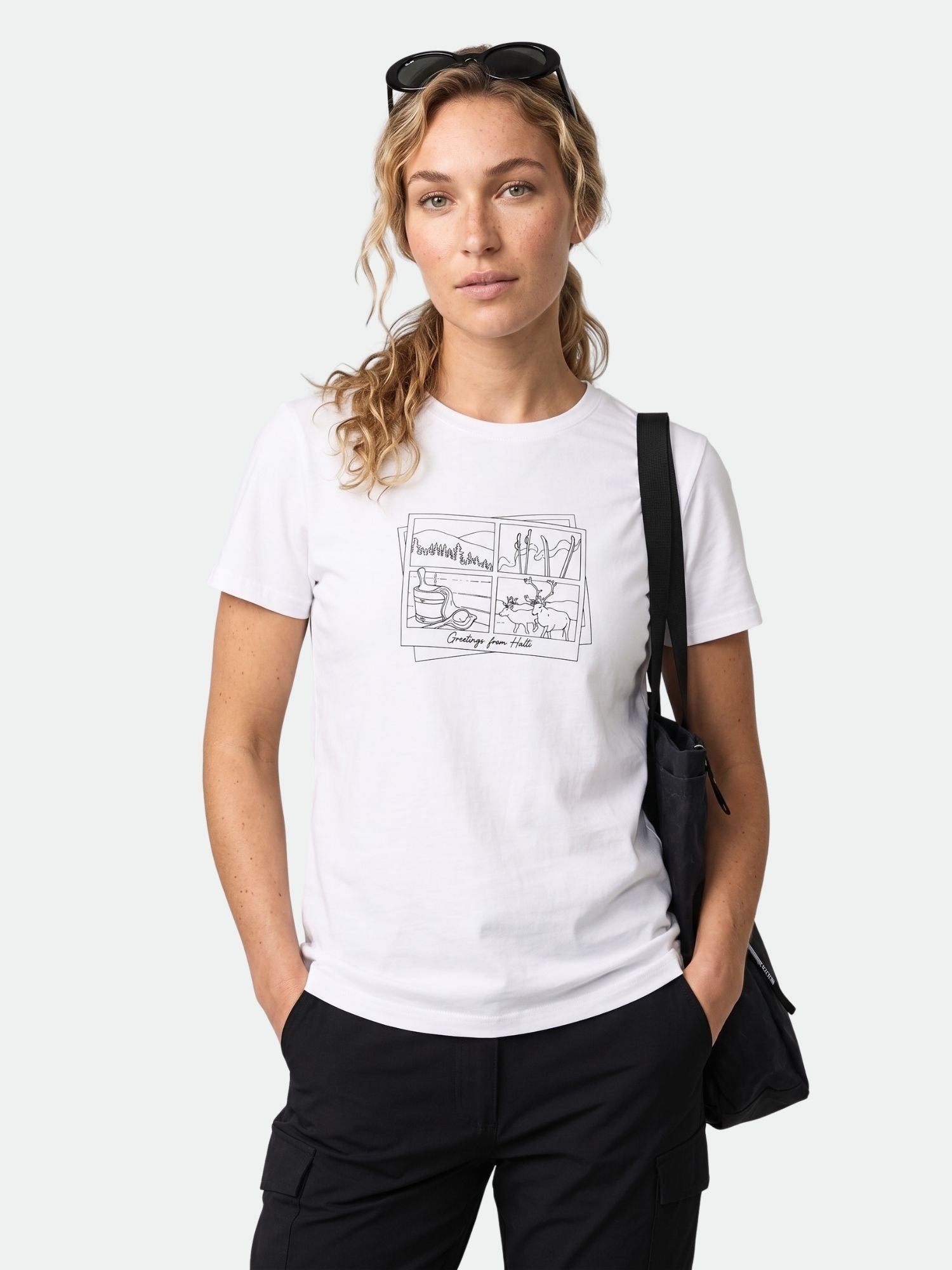 Woman in a white Halti Matka II T-shirt, black pants, and sunglasses, standing with one hand in her pocket.