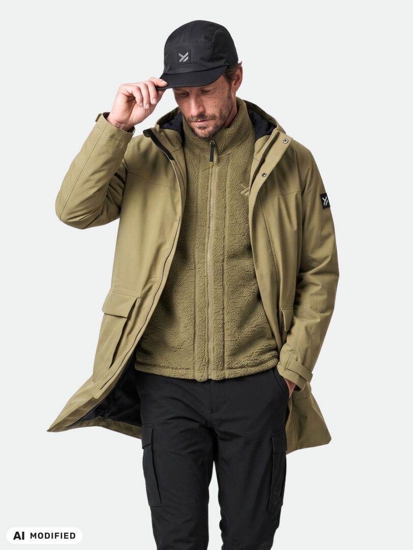 Man in Halti Kortteli DX Insulated Parka, fleece zip-up, black pants, and cap stands holding his cap with one hand.
