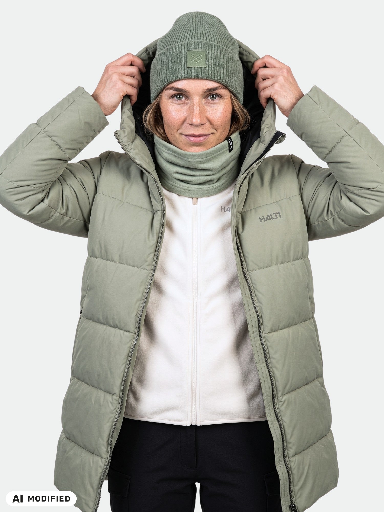 A woman in a green Halti Leinikko Puffer Parka, matching beanie, and neck warmer holds the hood with both hands.