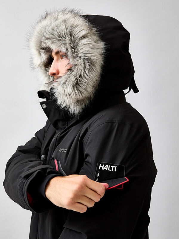 Person in a Halti 1976 Down Parka Unisex with a fur-lined hood adjusts a Halti patch on the sleeve.