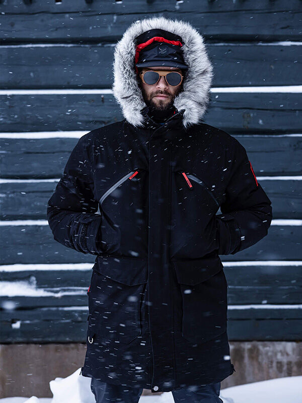 A person in a Halti 1976 Down Parka with fur hood, gloves, and sunglasses stands in falling snow by a wooden wall.