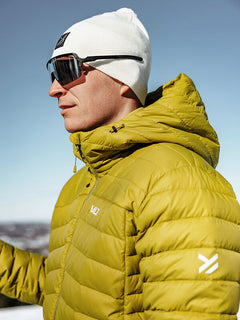 Man wearing Halti Huippu Down Jacket, white beanie, and sunglasses outdoors on a clear day.