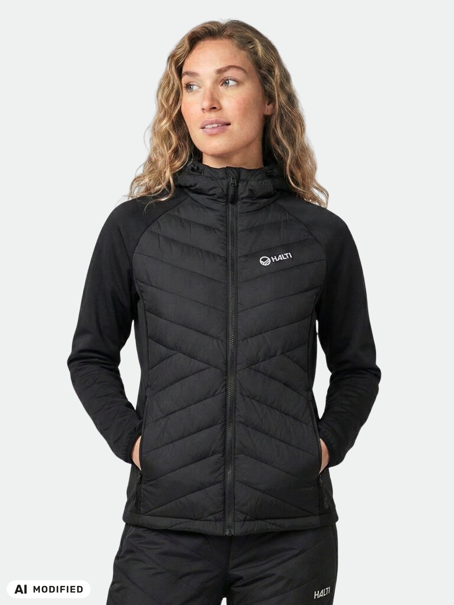 A woman wears the Halti Huippu Hybrid Down Jacket with a hood, hands in pockets, gazing slightly to the side.