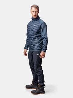 A man wears Haltis Huippu Hybrid Down Jacket, dark pants, and black hiking boots against a plain background.
