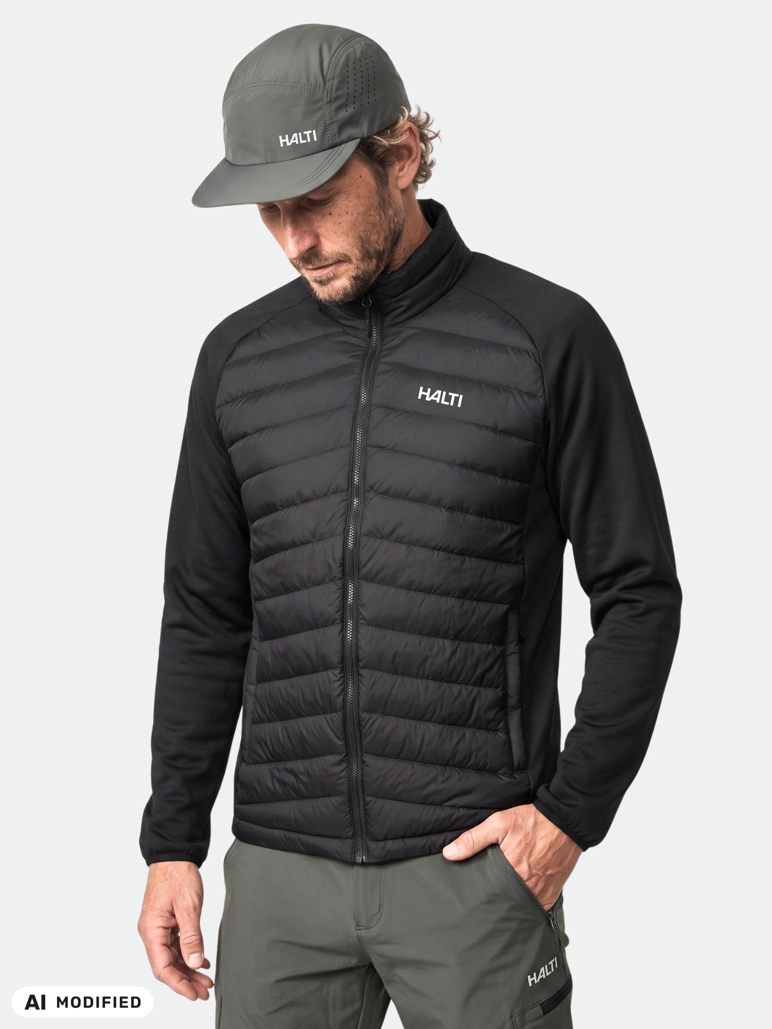 Man wears Halti Huippu Hybrid Down Jacket, gray cap and pants, standing against a plain light background.