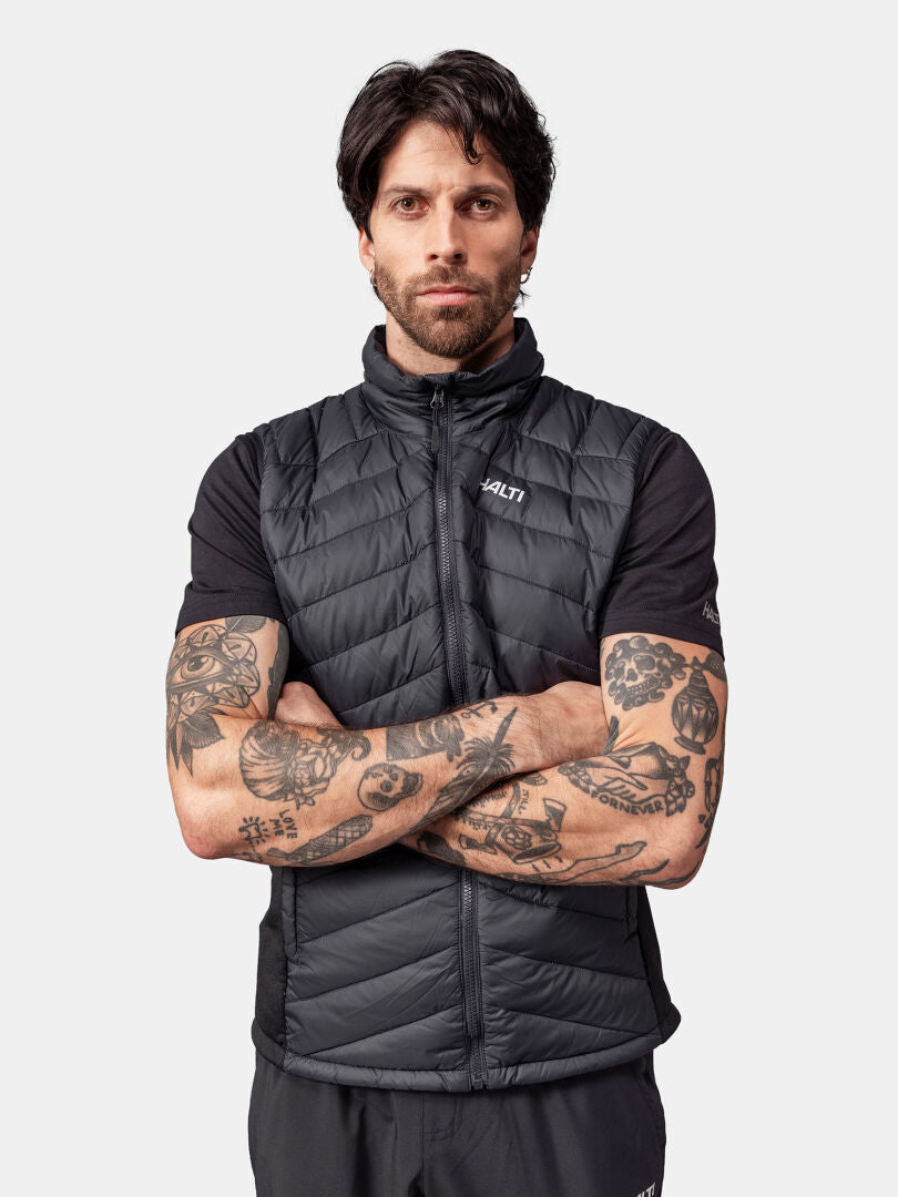 Tattooed man in Halti Huippu Down Vest, black with duck down insulation, stands with arms crossed.